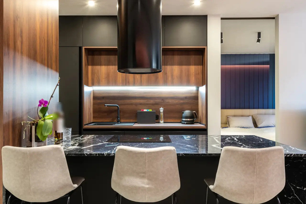 Cuisine d’appartement : îlot noir veiné, hotte cylindrique noire, crédence en bois foncé, chambre visible au fond.