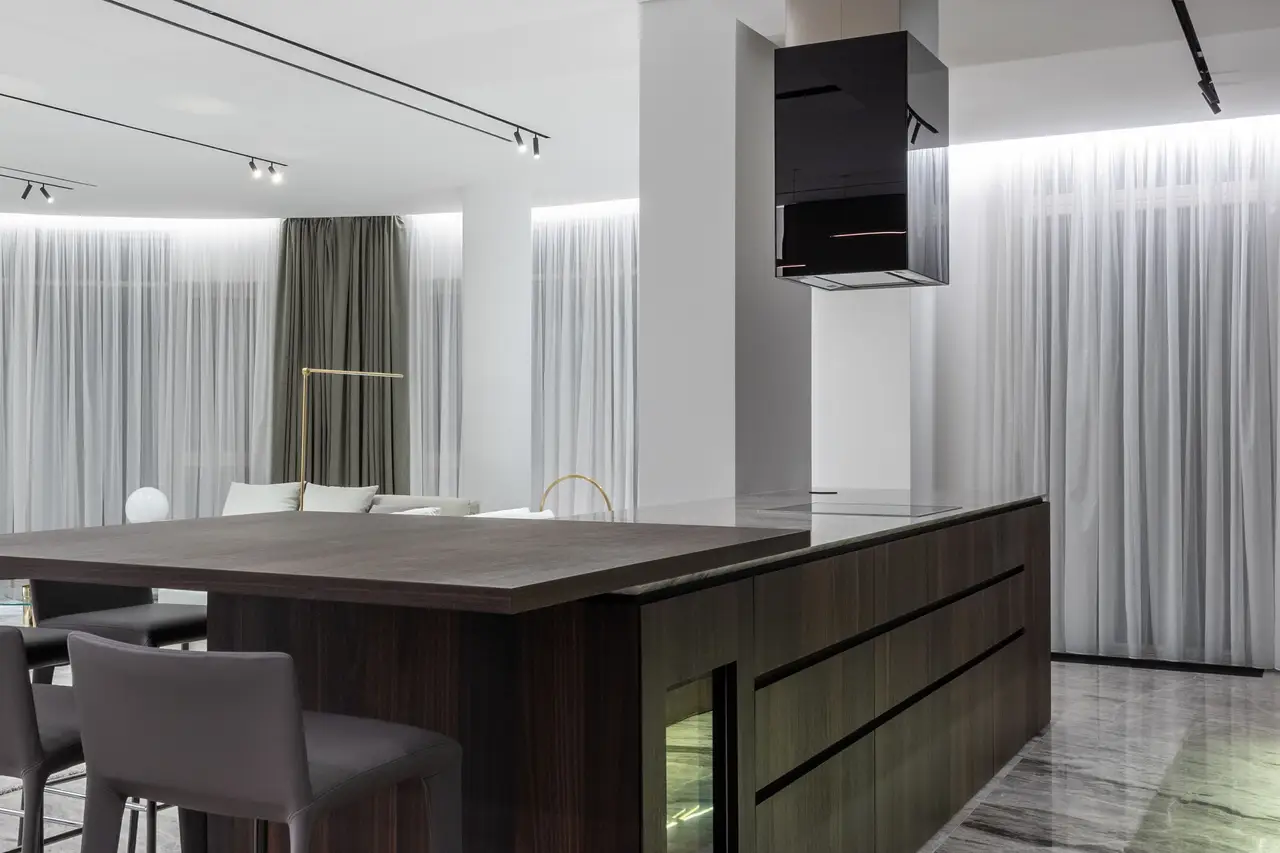Dark walnut island standing alone in a large pale room, curtains and stone floor, black suspended extractor.