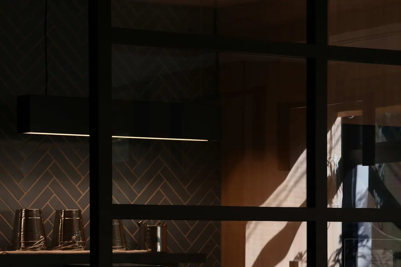 Verrière d’atelier devant une cuisine sombre, carrelage en chevrons et lumière de fin de journée sur le bois.